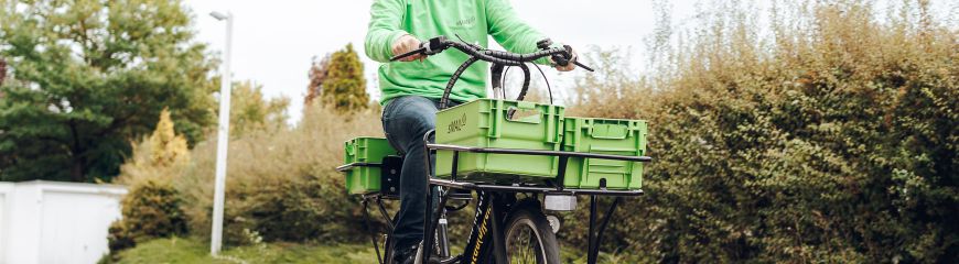 Ein Mann auf dem Fahrrad mit zwei grünen Kisten vorne an dem Fahrrad dran und der Mann hat einen grünen sMAIL Pullover an