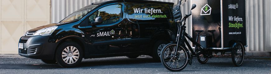 Ein schwarzes Auto (Caddy) mit dem sMAIL Logo und ein Fahrrad mit einer Box hinten drauf (Musketier) mit dem sMAIL Logo
