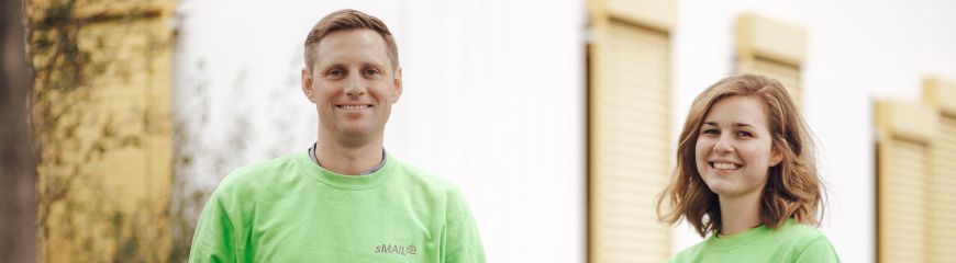 Eine Frau und ein Mann. Beide haben ein grünen sMAIL Pullover an und lächeln