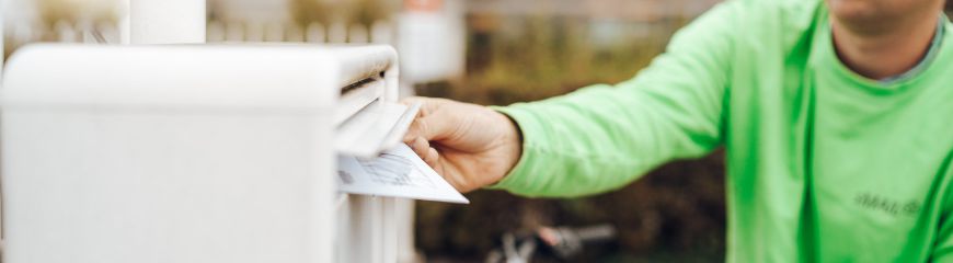 Ein Mann der einen grünen sMAIL Pullover an hat wirft breife in einen weißen Briefkasten