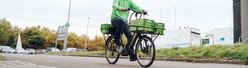 Ein Mann mit einem grünen sMAIL Pullover fährt Fahrrad und hat vorne am Fahrrad zweigrüne Kisten und auf dem Gepäckträger eine