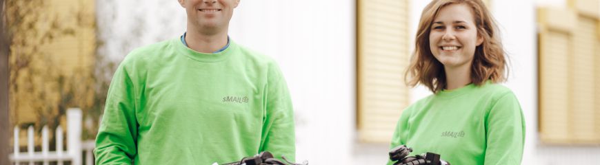 Eine Frau und ein mann stehen bei einem Fahrrad und haben grüne Kisten und Briefe in der Hand und auf dem Fahrrad