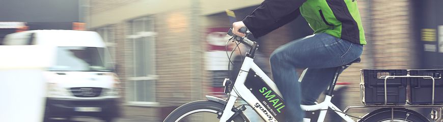 Ein Mann mit einer grünen sMAIL Jacke der gerade Fahrrad fährt und zwei schwarze Kiste auf dem Gepäckträger hat