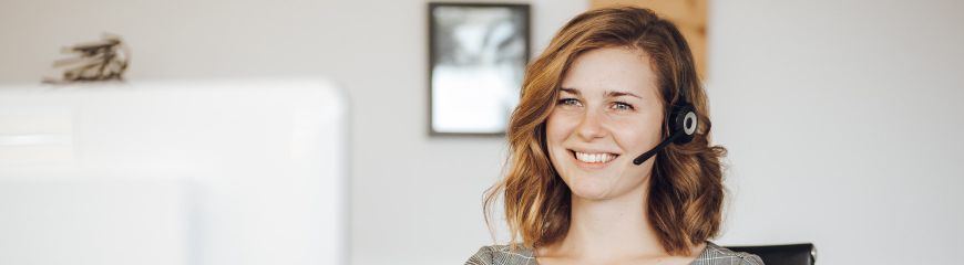 Eine Frau mit mittellangen Haaren die ein Headset an hat, am Computer sitzt und lächelt