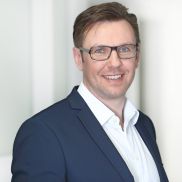 Ein man mit kurzen Haaren und einer Brille. Er hat ein weißes Hemd und ein dukelblaues Jackett an