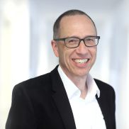 Ein Mann mit sehr kurzen Haaren und einer Brille. Er hat ein weißes HEmnd und ein shwarzes Jackett an