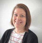 Eine Frau mit etwas kürzeren haaren. Sie hat ein weiß rot gesteiftes Tshirt und eine schwarze Strickjacke an